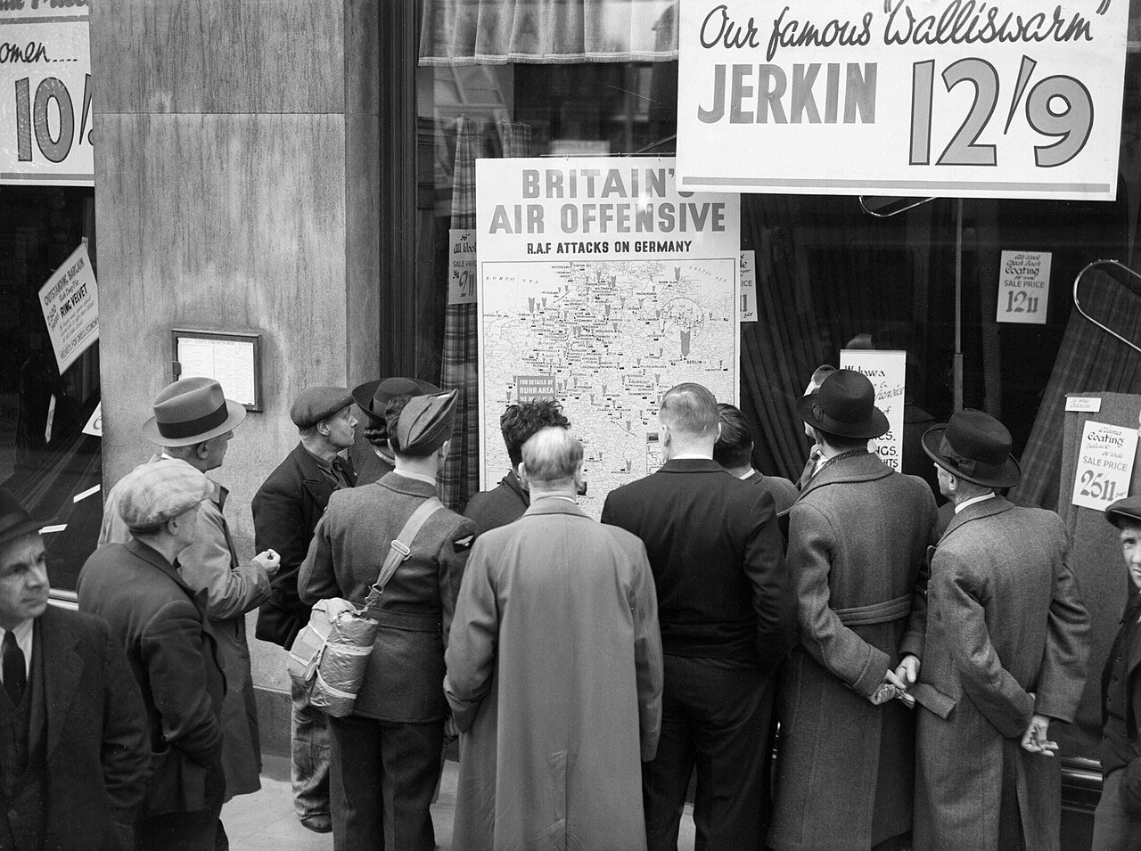 A British airman is amongst a group of civilians crowded around the window of a shop in Holborn, London, to look at a map illustrating how the RAF is striking back at Germany (1940) - Public Domain