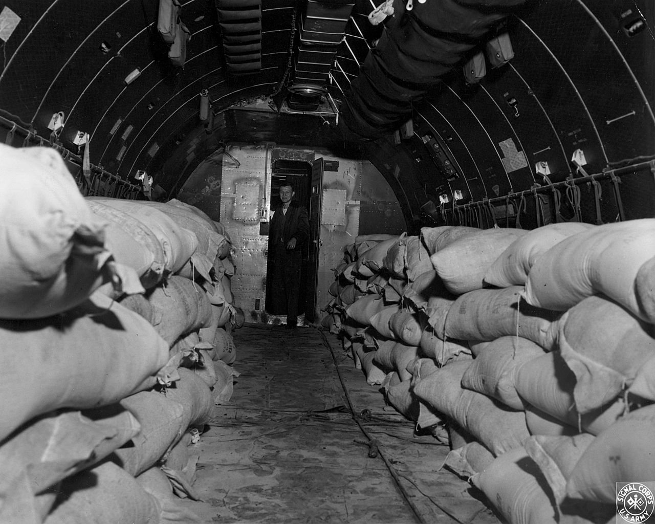 After loading of the C-54 air transport plane with sacks of flour by displaced Polish persons, German civilian personnel, and crewmembers of the plane prepares to take off from Rhine Main Airfield, Germany for Templehof Airbase in Berlin (1948) - Public Domain