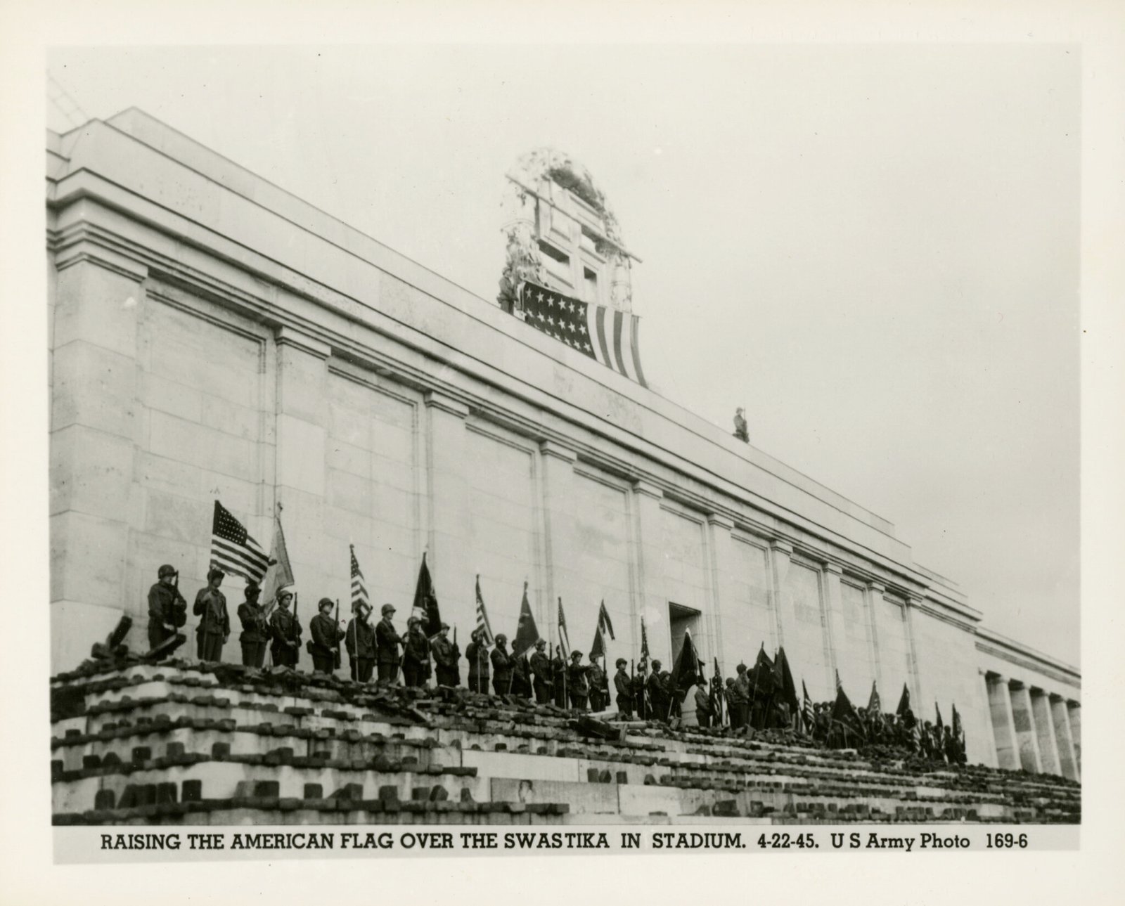 United States Army soldiers standing at attention in front of Deutsches Stadion as an American flag is raised over the swastika wreath on the top - April 22nd 1945