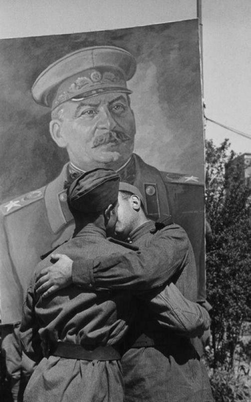 Two Red Arny soldiers embrace in a Socialist Kiss in front of an image of Joseph Stalin - Public Domain